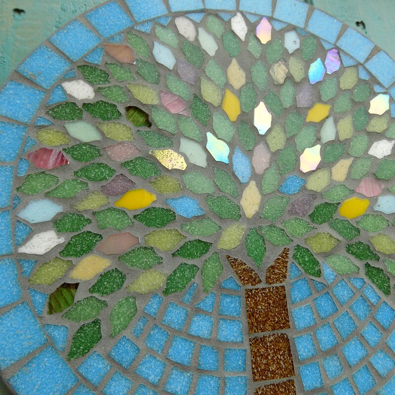 A mosaic garden plaque with a design of a spring tree with leaves made in the colours of spring blossom and flowers on a turquoise blue sky background