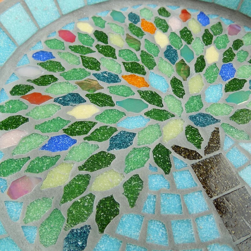 A mosaic garden bird bath with a design of a tree with the leaves in the colours of rich summer flowers and deep green leaves on a pastel sea green background