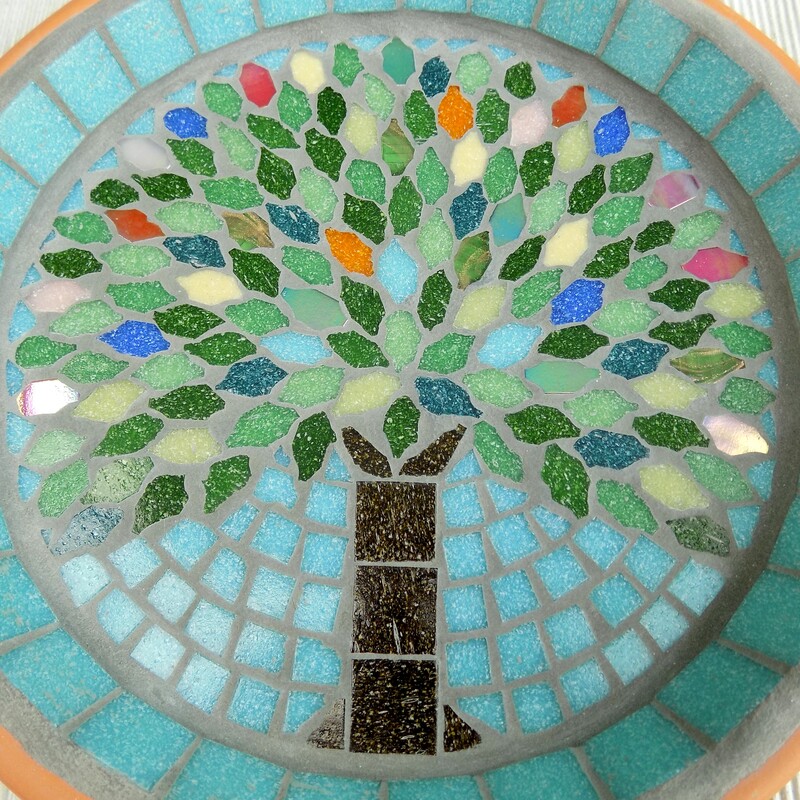 A mosaic bird bath with a design of a tree with the leaves in the colours of rich summer flowers and deep green leaves on a pastel sea green background