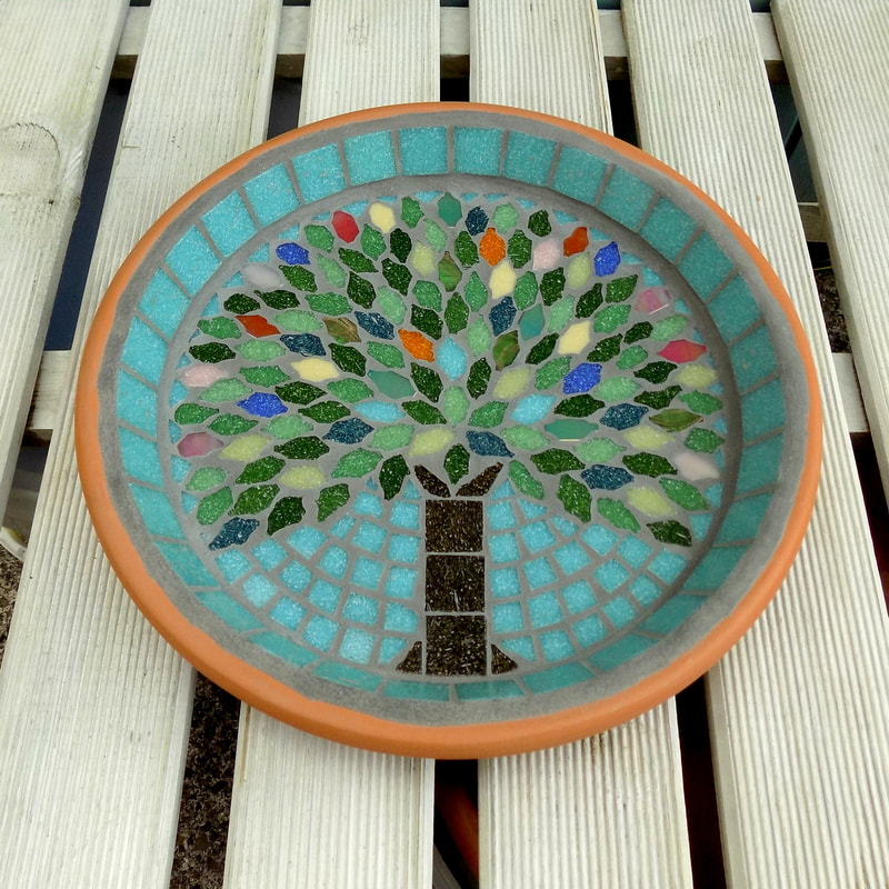 A mosaic birdbath with a design of a tree with the leaves in the colours of rich summer flowers and deep green leaves on a pastel sea green background