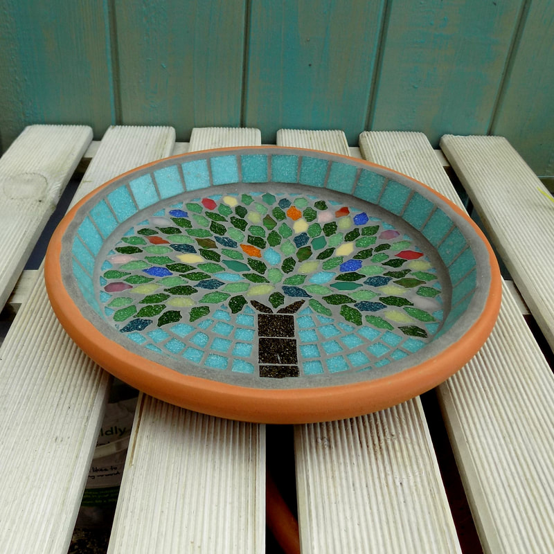 A mosaic garden birdbath with a design of a tree with the leaves in the colours of rich summer flowers and deep green leaves on a pastel sea green background