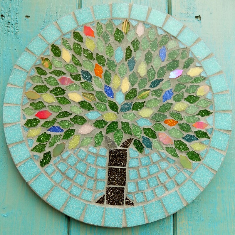 A mosaic hanging garden plaque with a design of a summer tree with leaves made in the colours of rich summer flowers and deep green foliage on a pastel sea green background