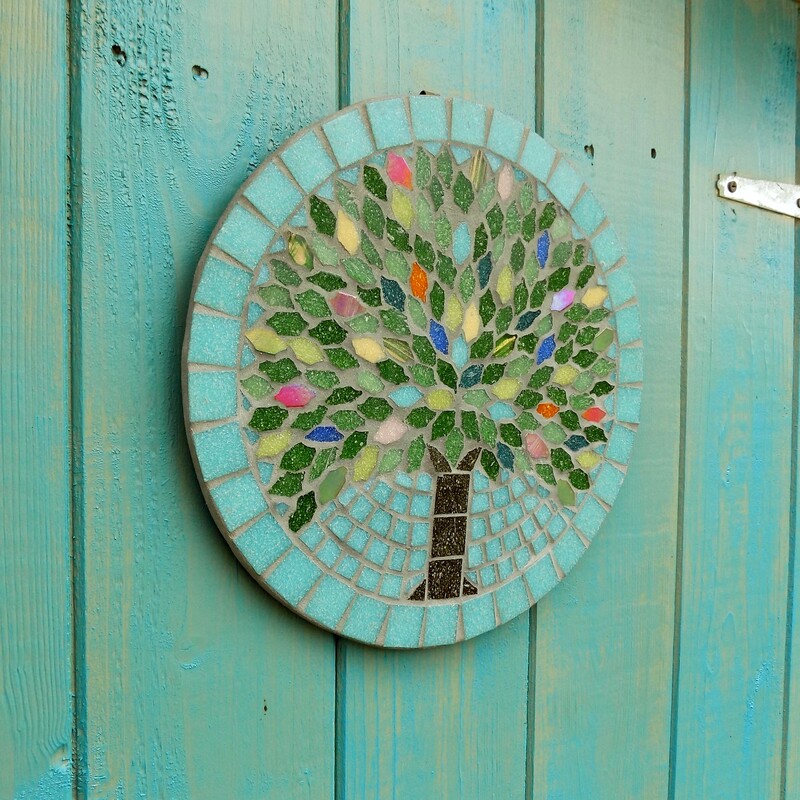 A mosaic garden plaque with a design of a summer tree with leaves made in the colours of rich summer flowers and deep green foliage on a pastel sea green background